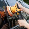 Corded orange and black dual action polisher with orange foam pad being held up against the side of a car by two hands.